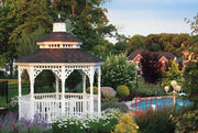 A wooden octagon Amish country gazebo, painted white with a shingled roof, sitting in a garden near a pool.  
