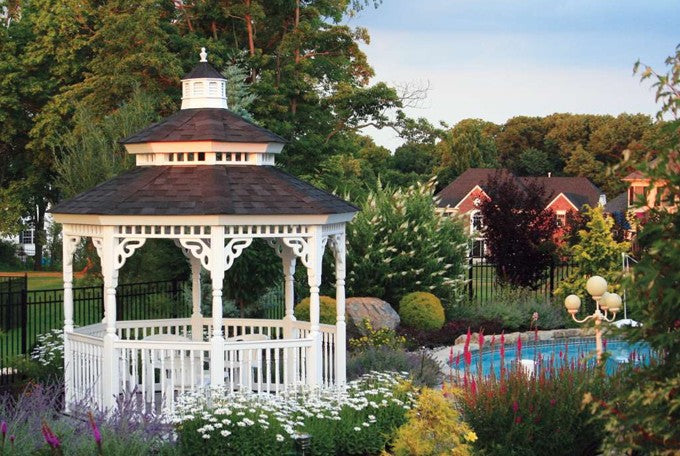 A wooden octagon Amish country gazebo, painted white with a shingled roof, sitting in a garden near a pool.  