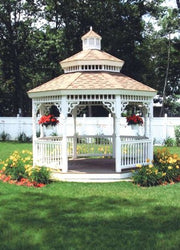 A white wooden gazebo with a brown shingled multi-tiered roof sits on a green lawn in a landscaped garden with flowers and a white fence in the background.