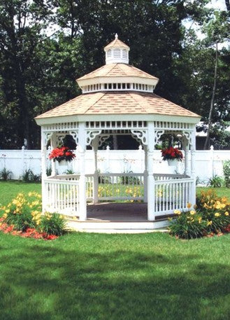 A white wooden gazebo with a brown shingled multi-tiered roof sits on a green lawn in a landscaped garden with flowers and a white fence in the background.