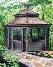 A wooden gazebo with a brown shingled roof and screen walls, situated on a stone patio surrounded by green trees and plants.