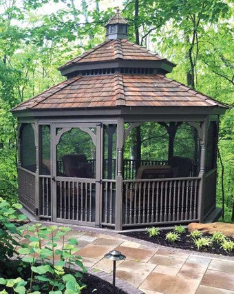 A wooden gazebo with a brown shingled roof and screen walls, situated on a stone patio surrounded by green trees and plants.