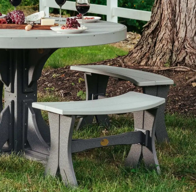 Outdoor patio dining set with a round gray table and two matching curved bench stools on grass beside a tree trunk.