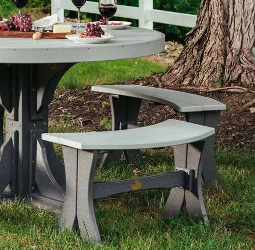 Outdoor patio dining set with a round gray table and two matching curved bench stools on grass beside a tree trunk.