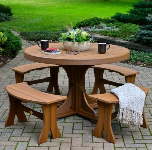 Round outdoor dining table with four matching curved bench seats, shown on a paved patio in a landscaped garden setting. The table is styled with a floral centerpiece, two black mugs, a small plate of fruit, and a white throw blanket draped over one bench.
