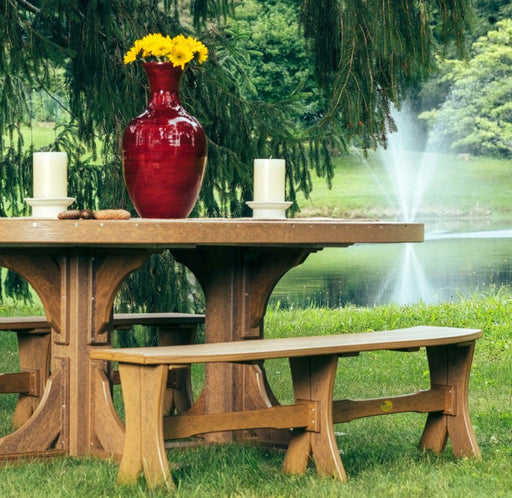 Outdoor picnic table and matching benches on a grassy lawn, with a red vase of yellow flowers and white candles on the tabletop, set against a pond with a fountain and trees.