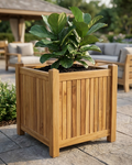 A square wooden planter box with a slatted front panel and sturdy corner posts, filled with dark soil and a large green leafy plant, set on a stone patio in an outdoor patio setting with lounge chairs and a pergola in the background.