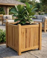 A square wooden planter box with a slatted front panel and sturdy corner posts, filled with dark soil and a large green leafy plant, set on a stone patio in an outdoor patio setting with lounge chairs and a pergola in the background.