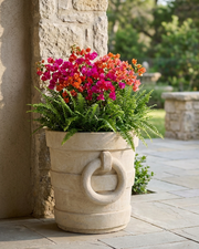 A large beige stone planter with a decorative ring handle, filled with green ferns and vibrant pink and orange flowers, set on a stone patio next to a stone pillar.