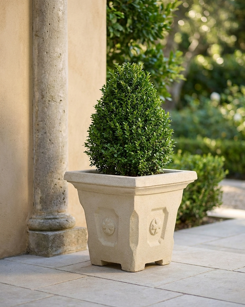 A square beige stone planter with decorative relief details, holding a neatly trimmed green topiary tree, set on a stone patio next to a stucco column in an outdoor garden setting.