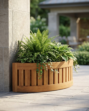 A curved wooden planter box with vertical slats, filled with green ferns and trailing ivy, placed beside a stone pillar on a paved patio.