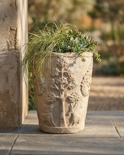 A tall, textured stone planter with carved tree and floral motifs, filled with green ornamental grass and succulents, set on a stone patio next to a stucco pillar.
