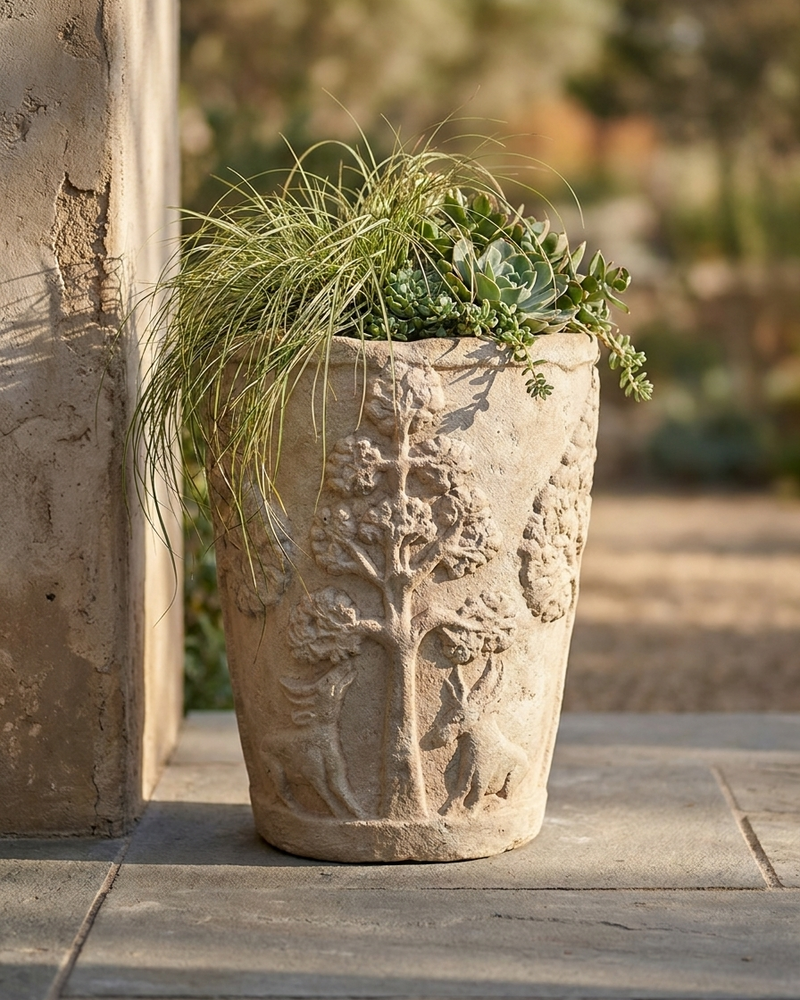 A tall, textured stone planter with carved tree and floral motifs, filled with green ornamental grass and succulents, set on a stone patio next to a stucco pillar.