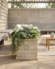 A large rectangular stone planter with a ring handle, filled with green ferns, white flowers, and purple lavender, placed on a stone patio in an outdoor patio setting.