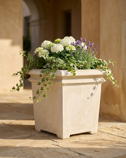  A Square limestone planter filled with white and purple flowers with greens palced on a stone patio.  