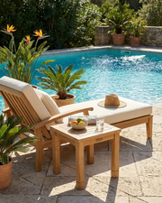 A wooden outdoor lounge chair with cream cushions sits beside a stone side table and pool, with a straw hat resting on the chair. The scene is set on a stone patio with potted plants and a swimming pool in the background.