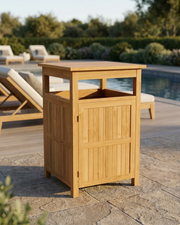 A light natural wood outdoor trash bin with a storage cabinet door, shown on a stone patio next to a swimming pool with lounge chairs in the background.