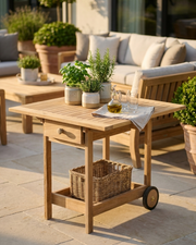 A light natural wood patio utility table with a single drawer, open lower shelf, and caster wheels, styled on a stone patio with potted plants and outdoor seating in the background.