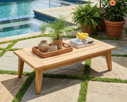 Toscana teak rectangular coffee table sitting on a stone and grass patio near a pool with plants.  