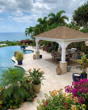 An outdoor patio and poolside pavilion scene with a tiled deck, gazebo, potted tropical plants, flowers, ocean view, and cloudy sky.