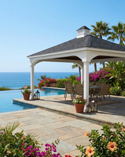 A white outdoor vinyl pavilion with a dark shingled roof sits beside a stone patio, pool, and ocean. The pavilion is furnished with a dining table and chairs, surrounded by tropical plants, palm trees, and bright pink flowers under a clear blue sky.