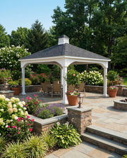 A white vinyl pavilion with a dark gray shingled roof sits on a stone patio in a landscaped garden with flower beds, potted plants, trees, and outdoor seating.