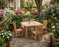 A 5-piece outdoor dining set made of light natural teak wood, including a rectangular table and four matching slatted chairs, arranged on a stone patio in a lush, garden-filled backyard setting.