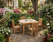 A 5-piece outdoor dining set made of light natural teak wood, including a rectangular table and four matching slatted chairs, arranged on a stone patio in a lush, garden-filled backyard setting.