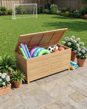 A wooden toy storage box with its lid open, filled with colorful pool toys, soccer balls, and basketballs, set on a stone patio in a backyard with a green lawn, potted plants, and a soccer goal in the background.