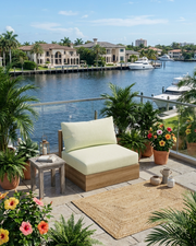 A modern outdoor lounge chair with light cream cushions and a natural wood frame sits on a stone patio overlooking a waterfront with boats and houses. The patio is decorated with potted palm trees, colorful flowers, a small side table, and a woven rug.