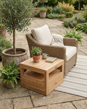 Teak side table next to a wicker chair on a rug on a stone patio with plants and trees.  