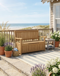 A wooden storage bench with armrests sits on a beachfront deck overlooking the ocean. The bench has a slatted back and seat, a cushionless storage base, and is styled with potted plants, a small side table, and a striped rug.