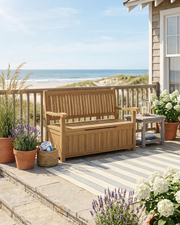 A wooden storage bench with armrests sits on a beachfront deck overlooking the ocean. The bench has a slatted back and seat, a cushionless storage base, and is styled with potted plants, a small side table, and a striped rug.