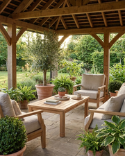 A wooden outdoor patio conversation set with three light beige cushioned armchairs and a matching rectangular coffee table, arranged under a rustic wooden pergola with potted plants and a garden background.