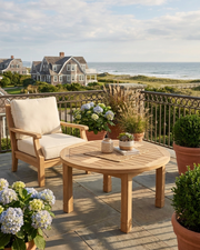 Outdoor patio furniture set on a stone terrace overlooking the ocean, featuring a round slatted wooden coffee table and a matching wooden armchair with cream cushions, styled with potted plants and a railing.