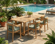 Outdoor dining set with a rectangular wooden table and six matching armchairs with light green cushions, arranged on a stone patio beside a swimming pool and tropical plants.